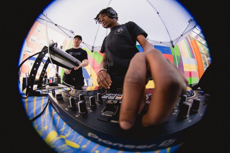 A fisheye-type photograph of two DJs mixing music at a turntable under a tent with colorful decorations; one is focused on adjusting the equipment while the other observes.