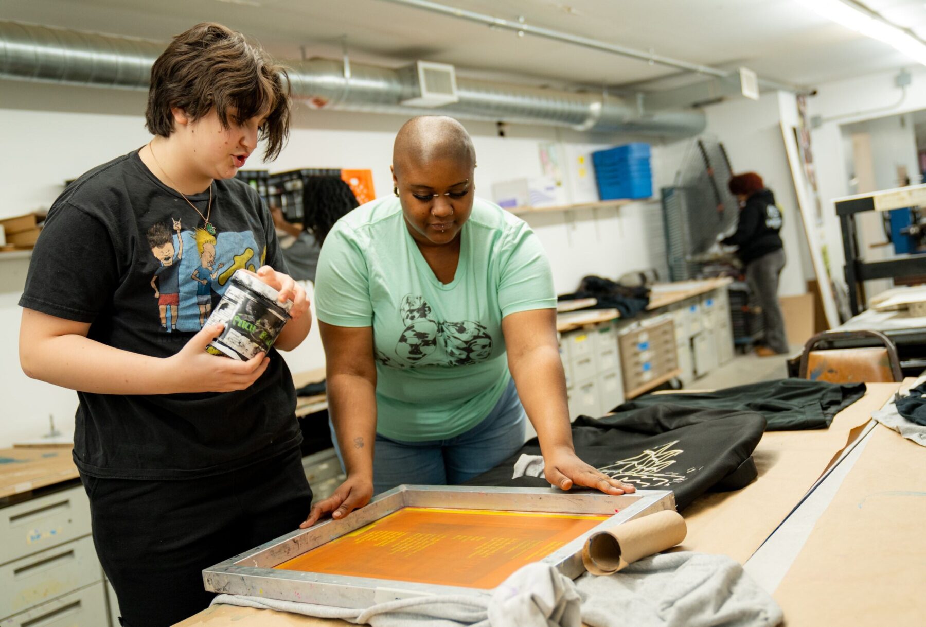 Two people in the process of silk screen printing on a sweatshirt. One person holds a container of ink while the second person holds the silk screening frame.