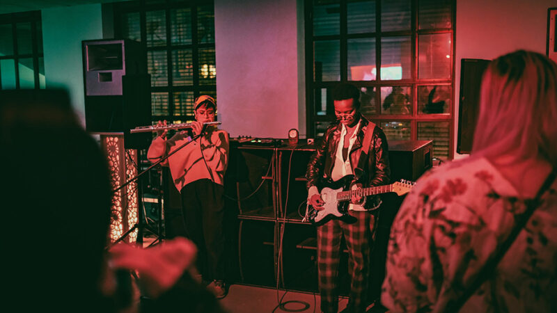 Musicians performing at a live concert, with one playing the guitar and the other using a synthesizer, in front of an audience in a dimly lit venue with windows in the background.