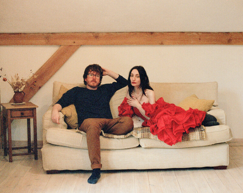 A person in a red outfit sitting on a sofa next to a person in brown pants and a navy shirt, in a room with wooden beams and minimal decor.