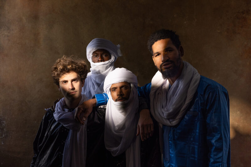 Four individuals wearing traditional Tuareg attire pose against a textured brown background.