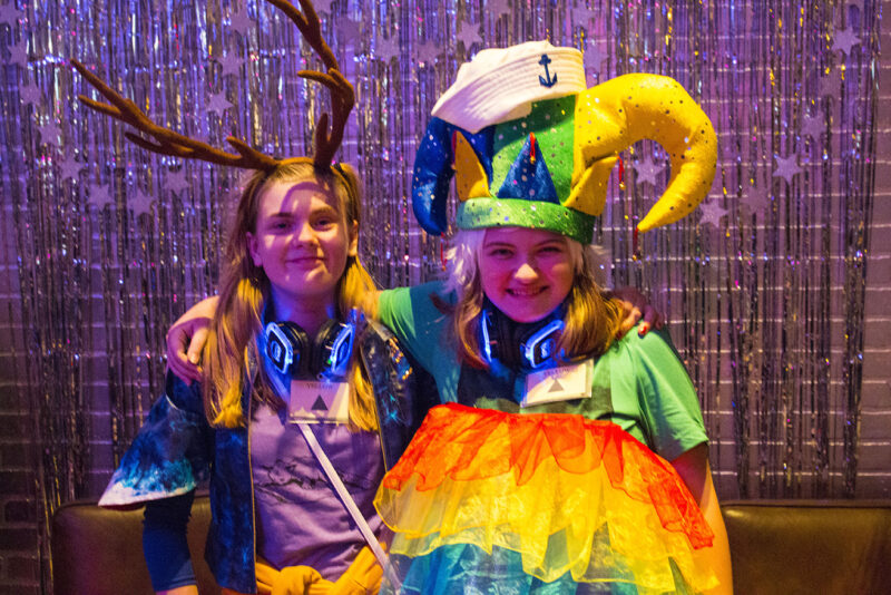 Two individuals in colorful costumes standing in front of a shimmering backdrop. One person is dressed with a large jester hat with a sailors had on top, and the other has antlers. Both are smiling.