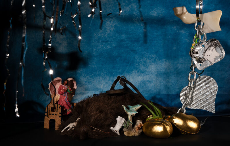 A whimsical still life display featuring a variety of objects including a dolphin statue, plush toy, golden plush balls, an angel statue, a silver heart keychain, and a cartoon character-like star, set against a textured blue background adorned with twinkling tinsel.