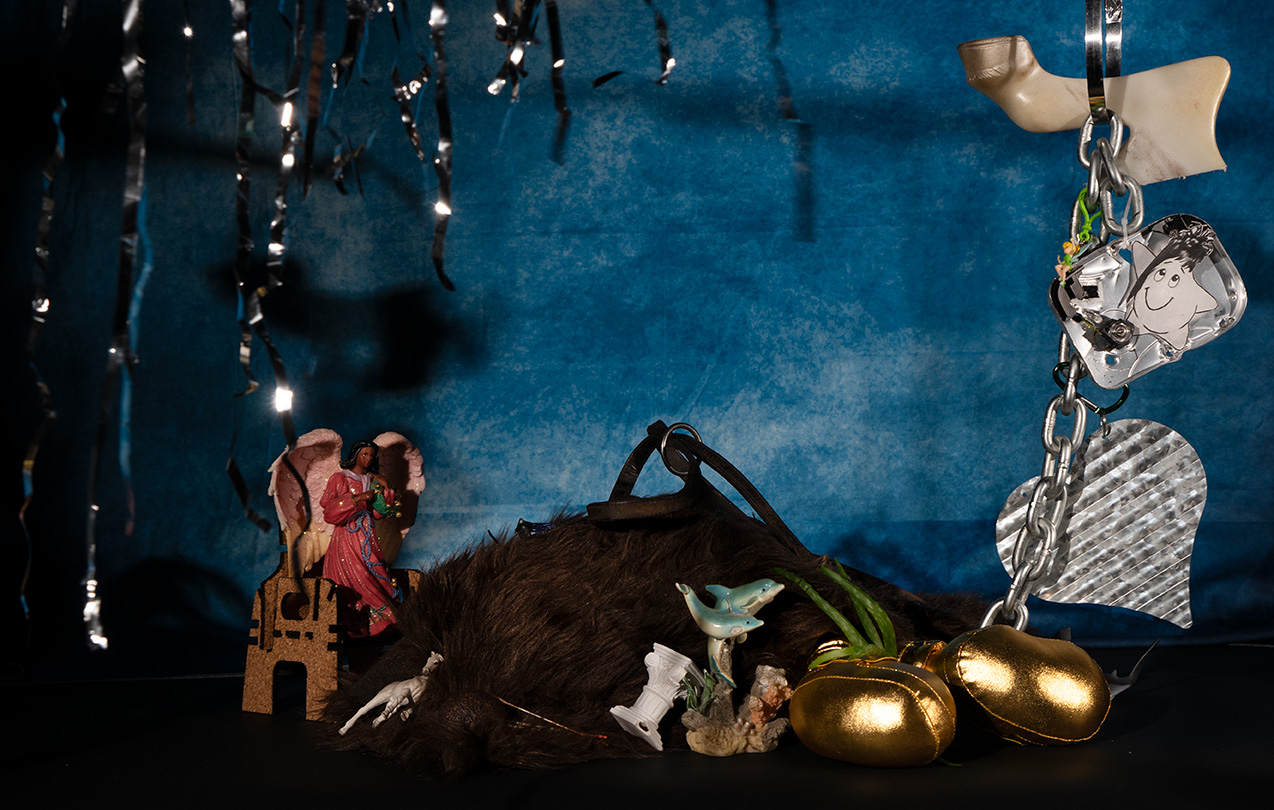 A whimsical still life display featuring a variety of objects including a dolphin statue, plush toy, golden plush balls, an angel statue, a silver heart keychain, and a cartoon character-like star, set against a textured blue background adorned with twinkling tinsel.