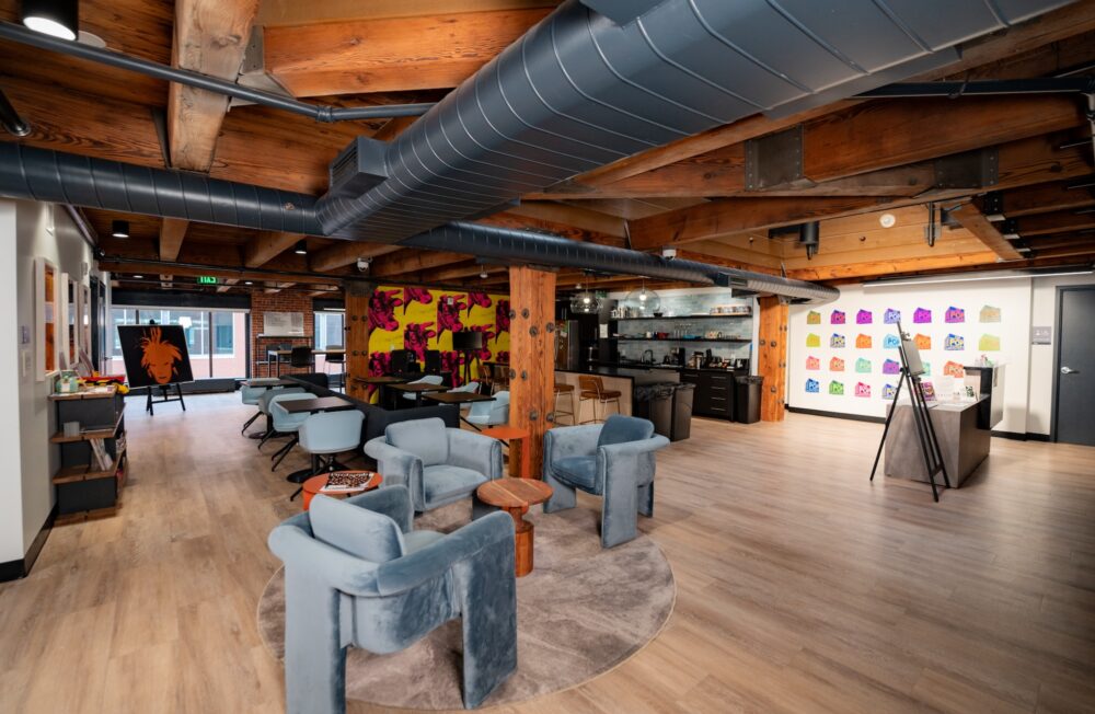 An open floor plan room with exposed duct work hanging from the ceiling. A kitchen counter, island, and refrigerator is in the center of the room. The rest of the room has a variety of chairs and small tables.