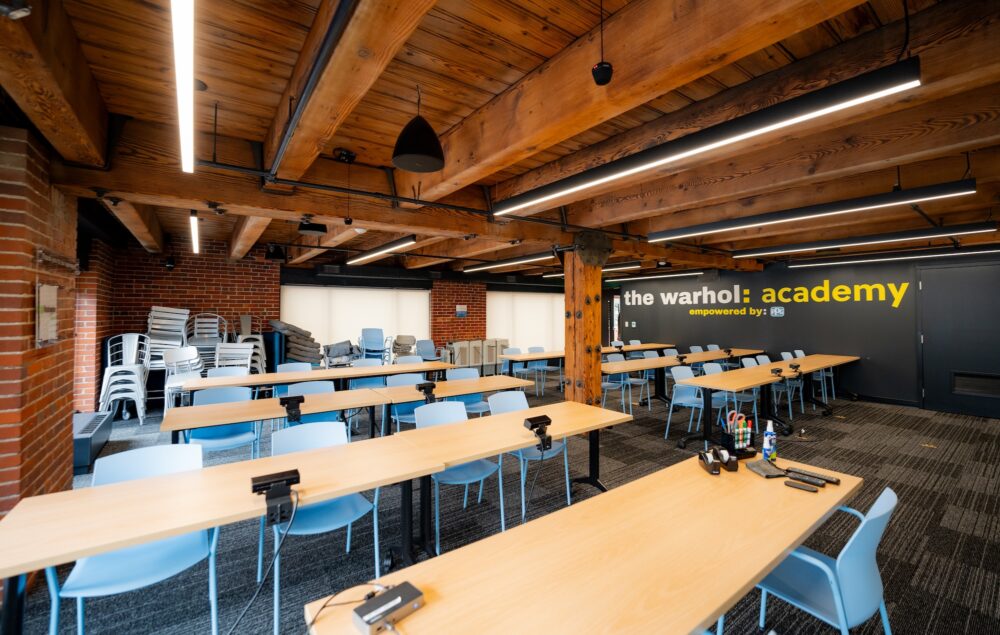 A large room with six long tables, all with light blue chairs at them. Several stacks of chairs are in the back of the room. One wall is painted black and has the words the warhol: acadmey printed on it.