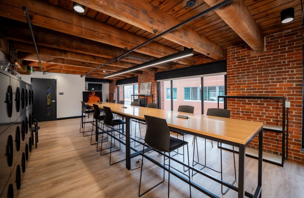 A room with an exposed brick wall, and wooden ceiling. In the room is a long thin table with 9 empty chairs.