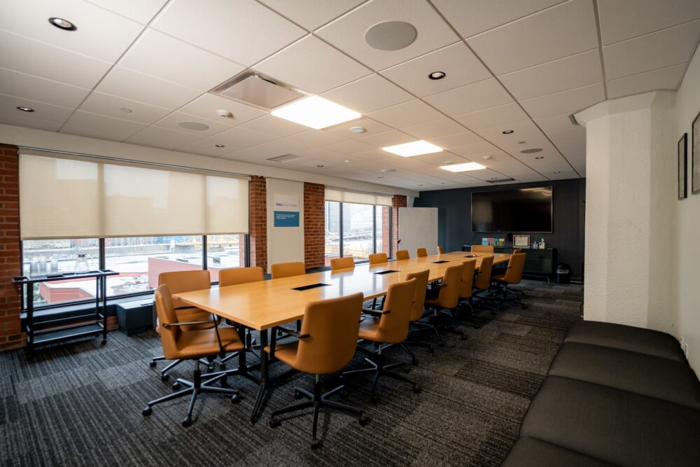 A long conference room. A long table with chairs takes up most of the center of the room. Two large floor to ceiling windows overlook a city. One large television is mounted on the wall.