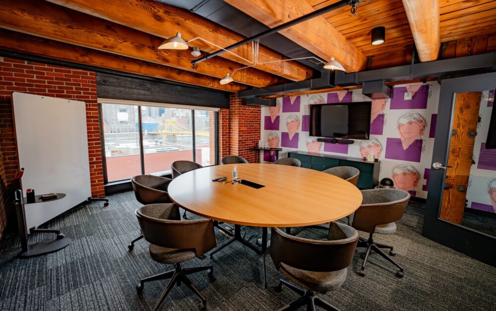 A room with a large round wooden table in the center. Eight empty chairs are at the table. A large floor to ceiling window overlooks a river with a bridge. The one wall has colorful wallpaper and a single television mounted in the center of the wall.