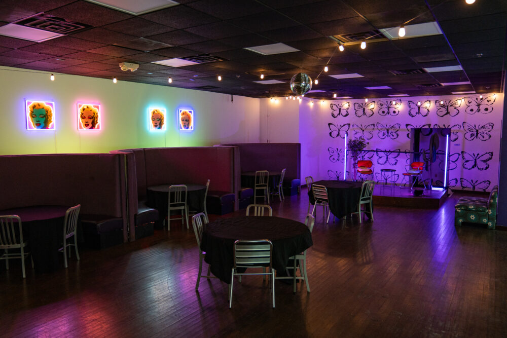 A room with 4 round tables that each have silver chairs. The left wall has 4 portraits of Marilyn Monroe by Andy Warhol with neon lights surrounding them. The right wall is white with black drawings of butterflies.