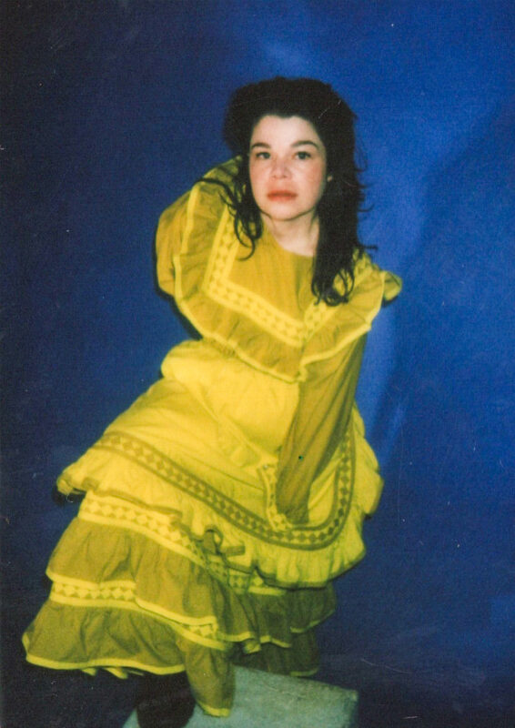 Samantha Crain is in a vibrant yellow, tiered dress posing against a blue background.