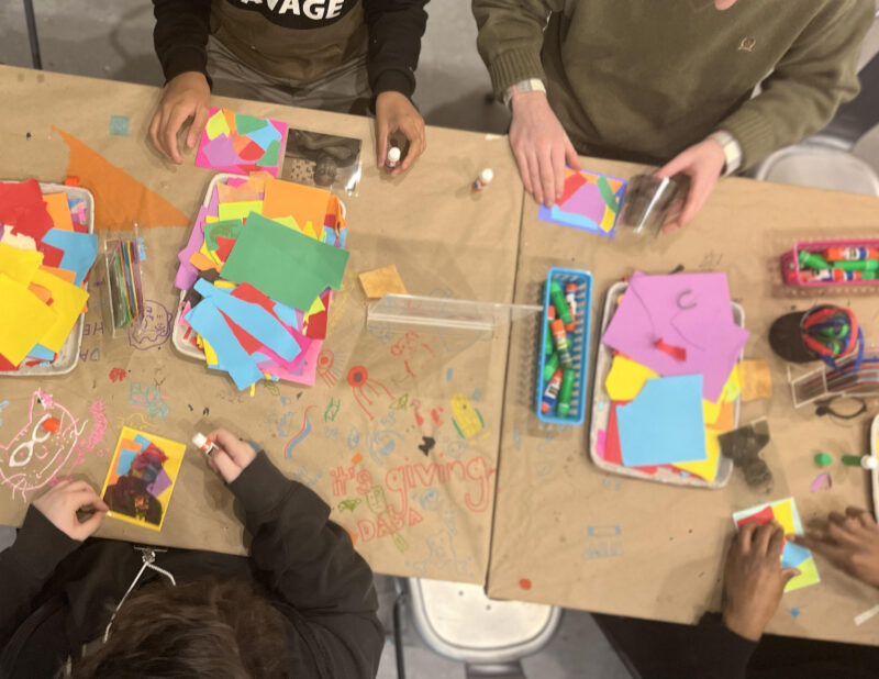 Overhead view of several individuals engaged in crafting with colorful paper and markers at a table covered in drawings and art supplies.