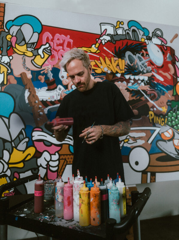 Person standing in front of a colorful graffiti wall, looking downward with a table of paint supplies in the foreground.