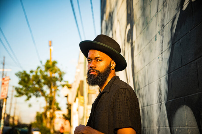 A person standing against a sunlit building wall, wearing a black hat and looking confidently at the camera.