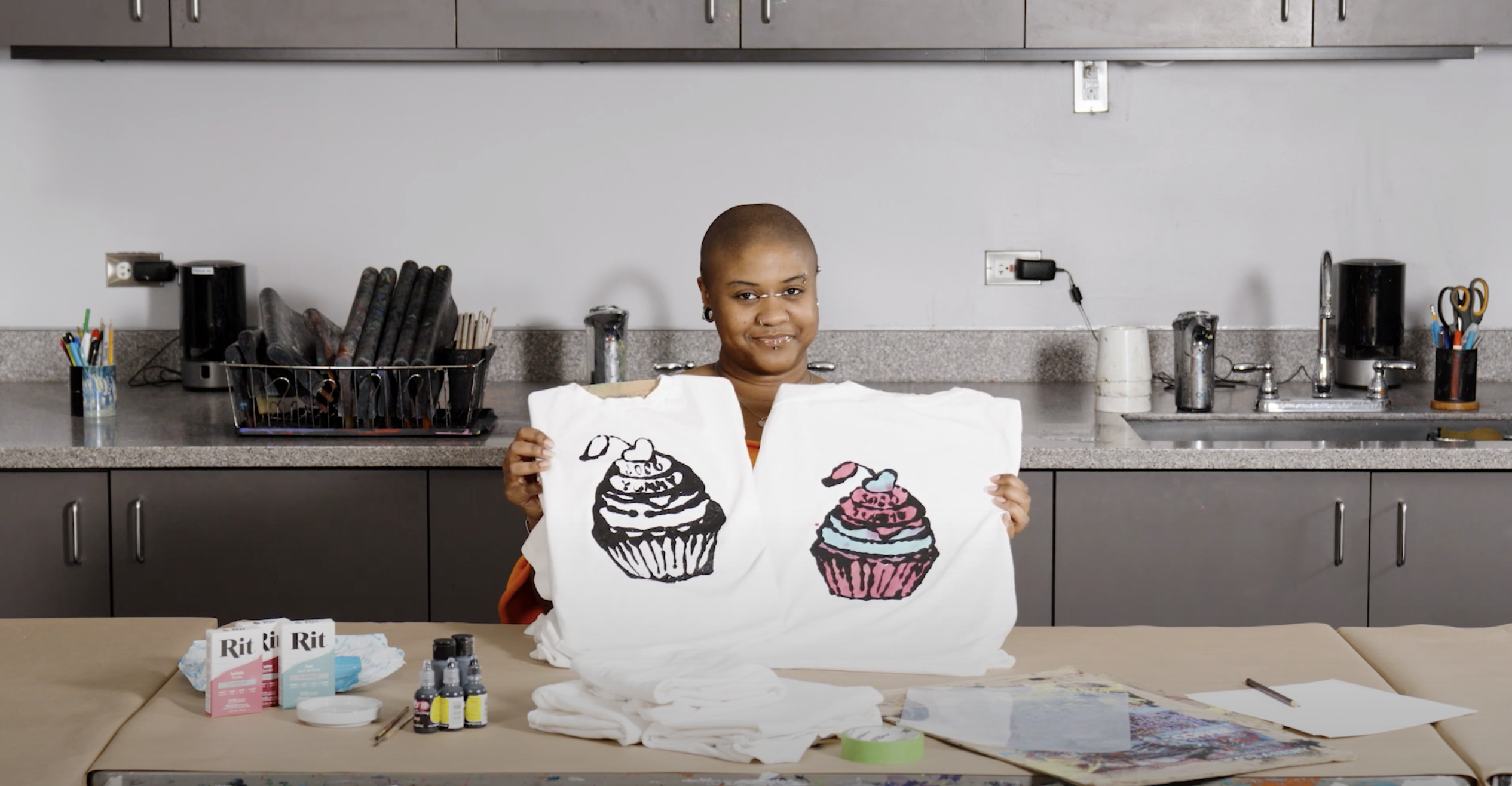 A person sits at a table that holds art supplieds. The person is holding two tshirts, both decorated with a drawing of a cupcake. The right shirt has colored the cupcake pink and blue.