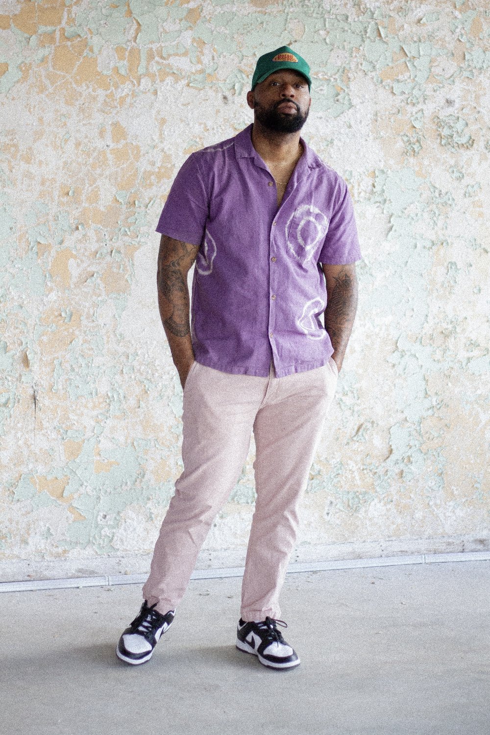 A person standing against a rustic wall, wearing a purple shirt, beige pants, and black and white sneakers.