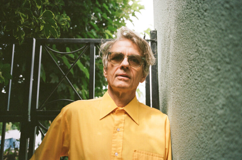 A person in a bright yellow shirt and silver-framed glasses stands between a black gate and a textured wall, looking into the camera under a canopy of green foliage.